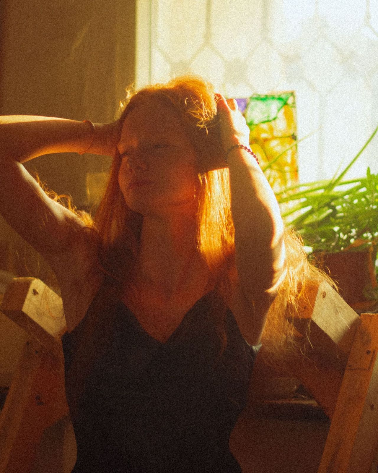 A woman with long copper hair, hands in her hair, in warm window light with plants behind her